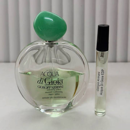A bottle of Giorgio Armani Acqua di Giola Eau de Parfum next to a small travel perfume sample vial on a white surface with a white curtain background.