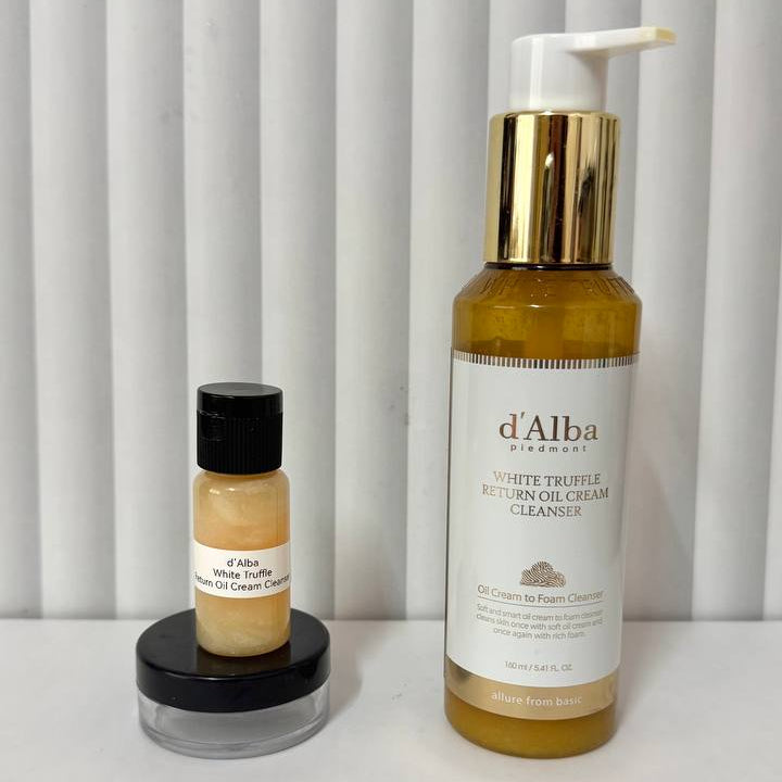 Two d'Alba Cosmetics skincare products on a white surface against a white slatted background: a larger amber bottle with a white pump and gold collar labeled White Truffle Hydrating Cleanser, and a smaller amber bottle with a black cap on a black lid labeled White Truffle Hydrating Balm.