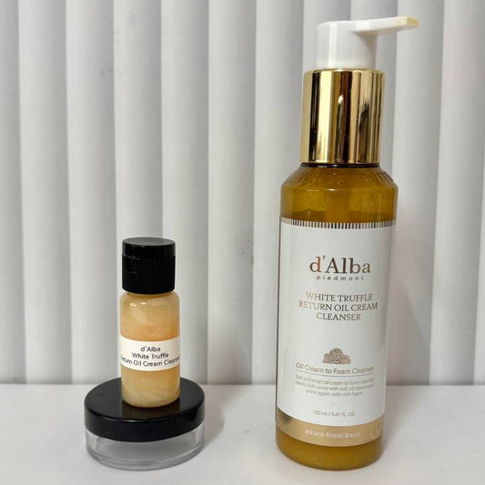 Two d'Alba Cosmetics skincare products on a white surface against a white slatted background: a larger amber bottle with a white pump and gold collar labeled White Truffle Hydrating Cleanser, and a smaller amber bottle with a black cap on a black lid labeled White Truffle Hydrating Balm.